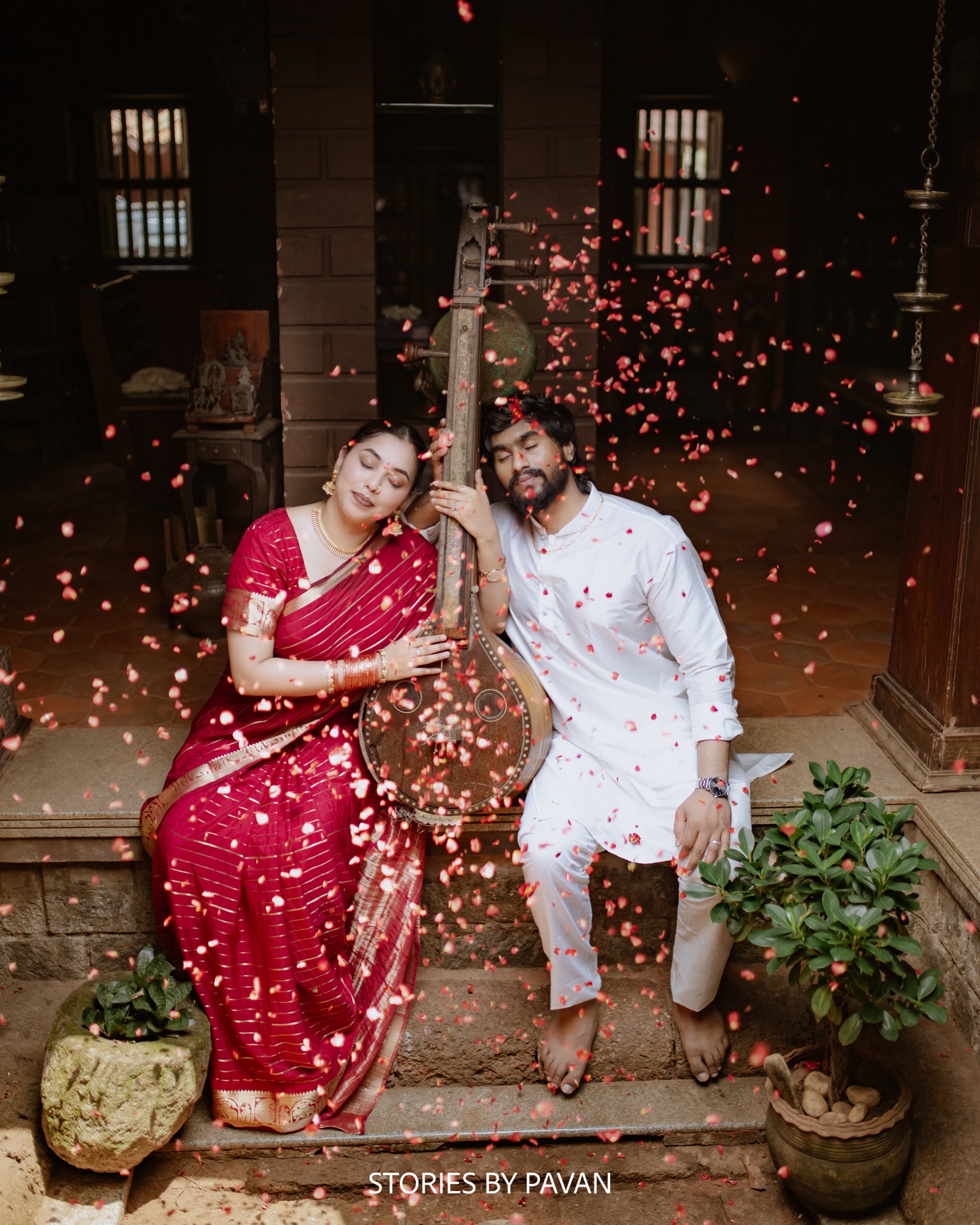 Bride and groom seated together on a traditional Kerala courtyard step, holding a tanpura as rose petals fall around them