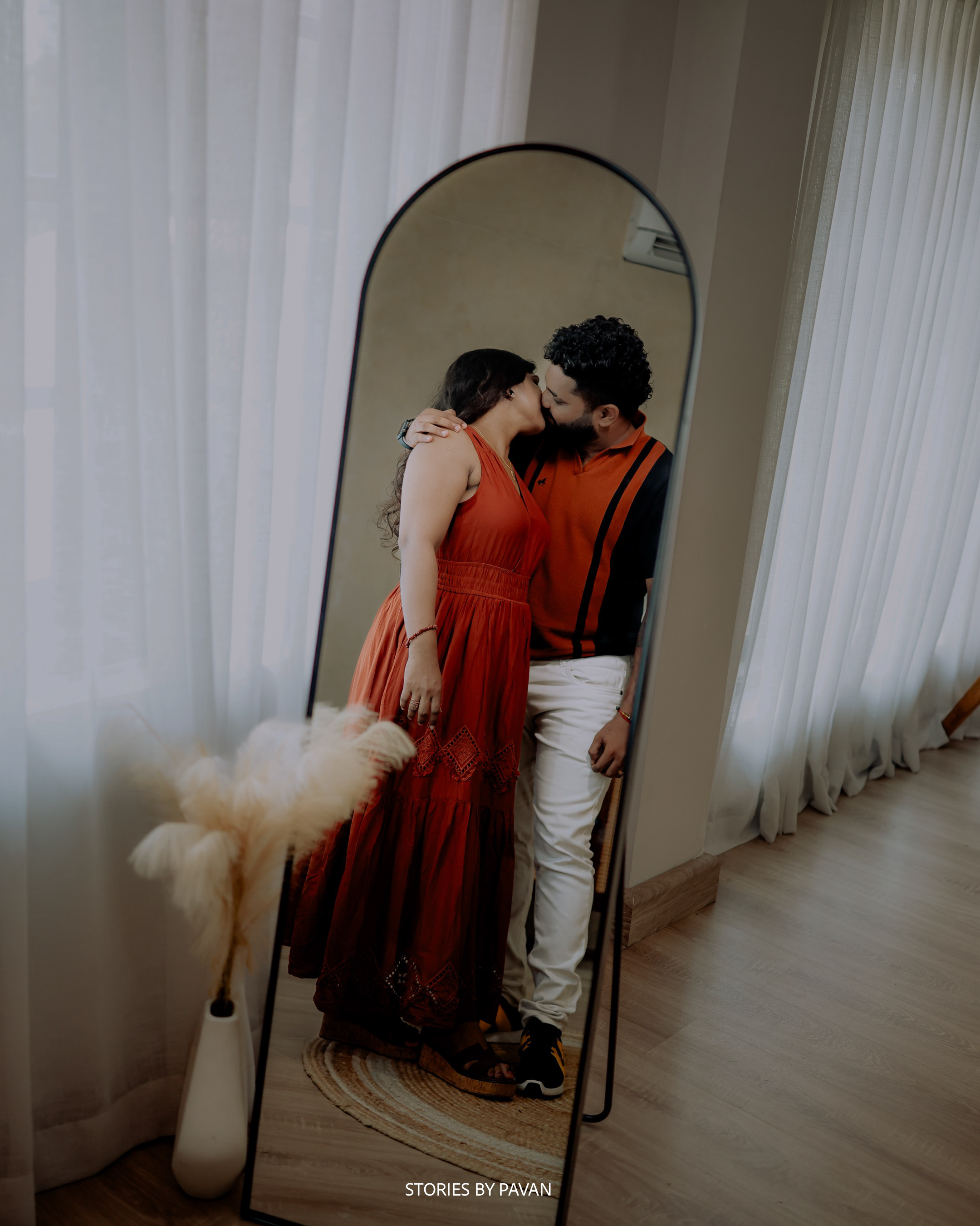 Couple sharing a kiss reflected in a tall mirror, softly lit by sheer curtains