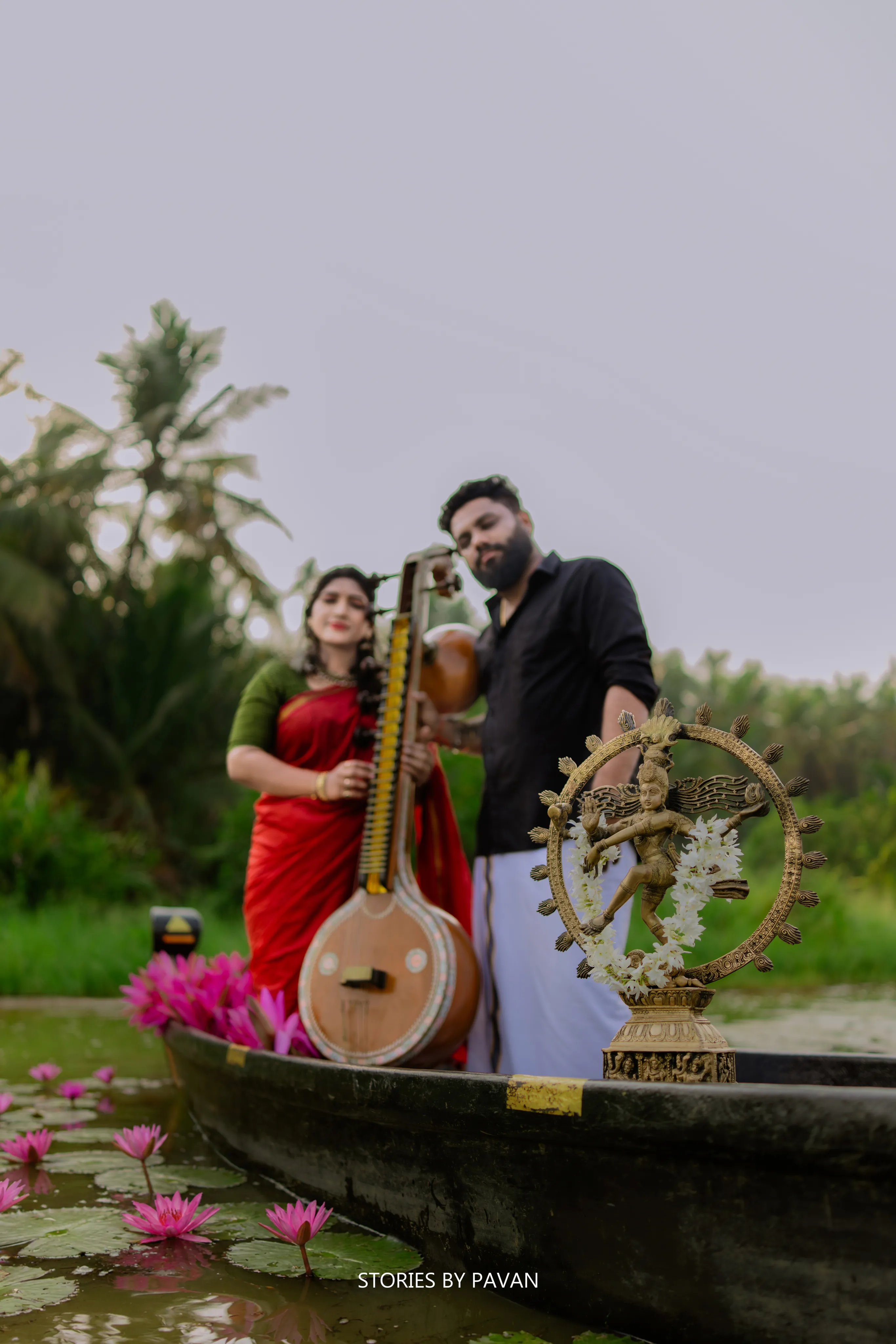 Couple standing in a wooden boat among pink lotus flowers, holding a tanpura and a small Nataraja idol