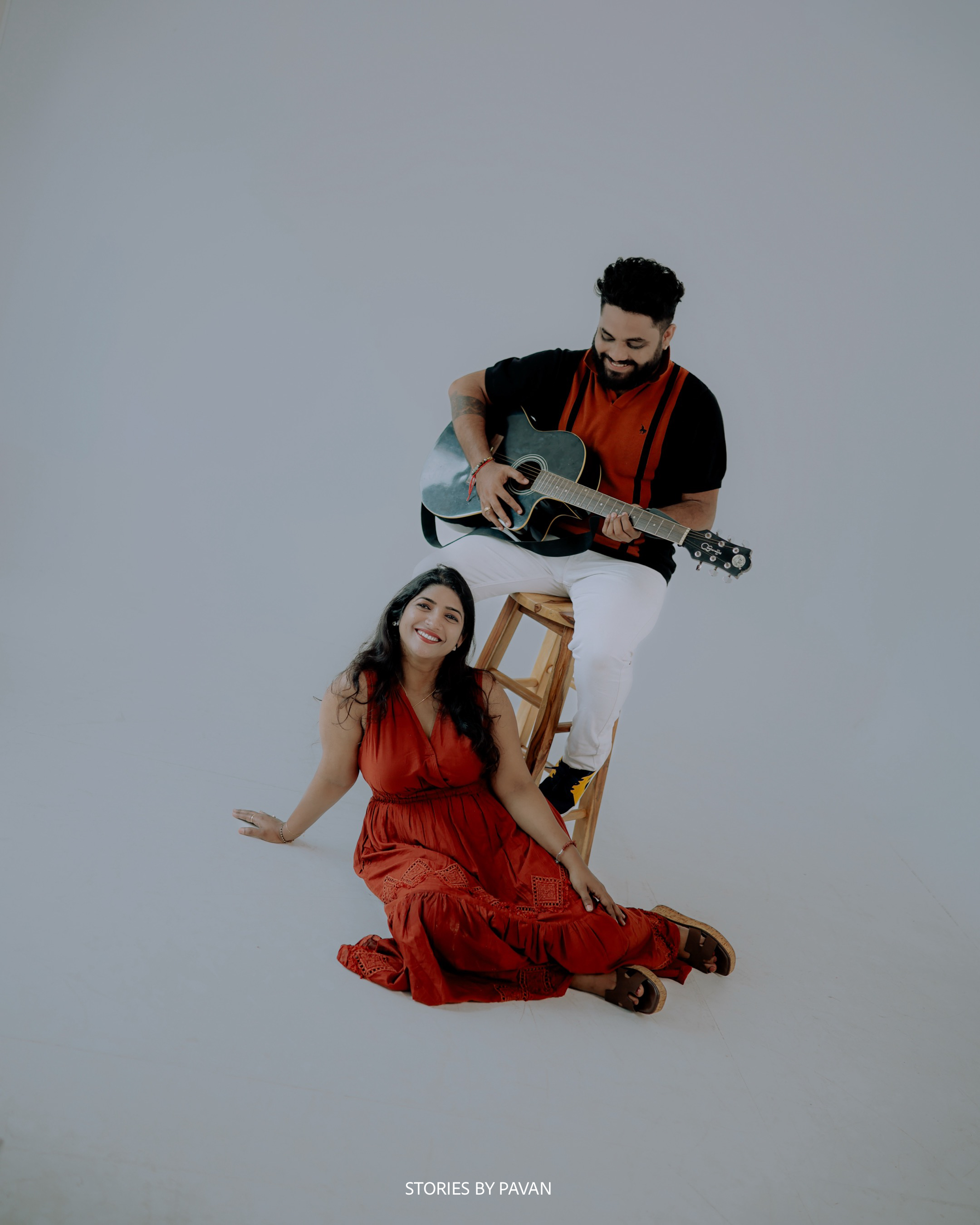 Groom playing guitar on a stool while the bride sits smiling on the floor beside him