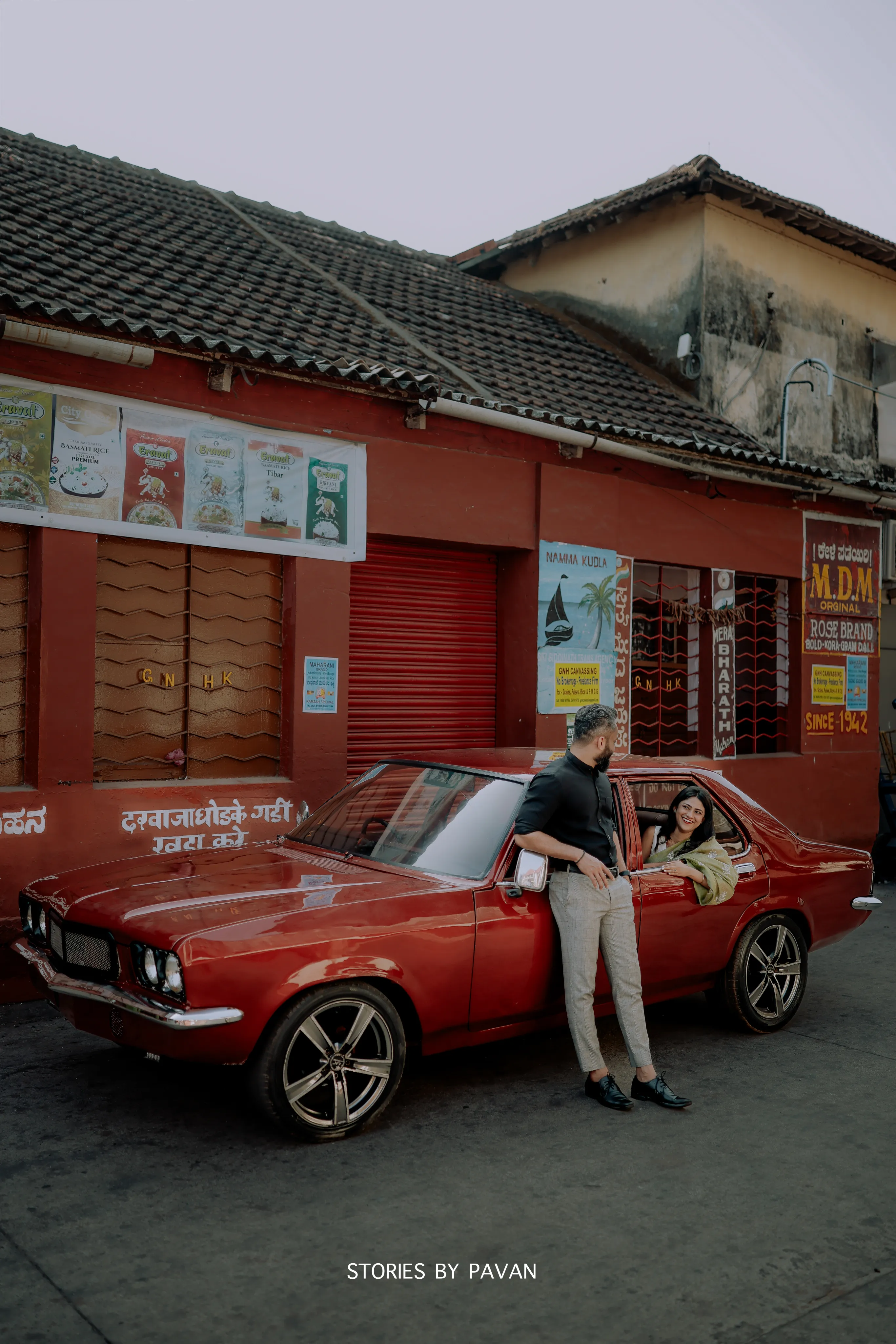 Couple by a vintage red car outside an old red-painted shopfront on a Mangalore street