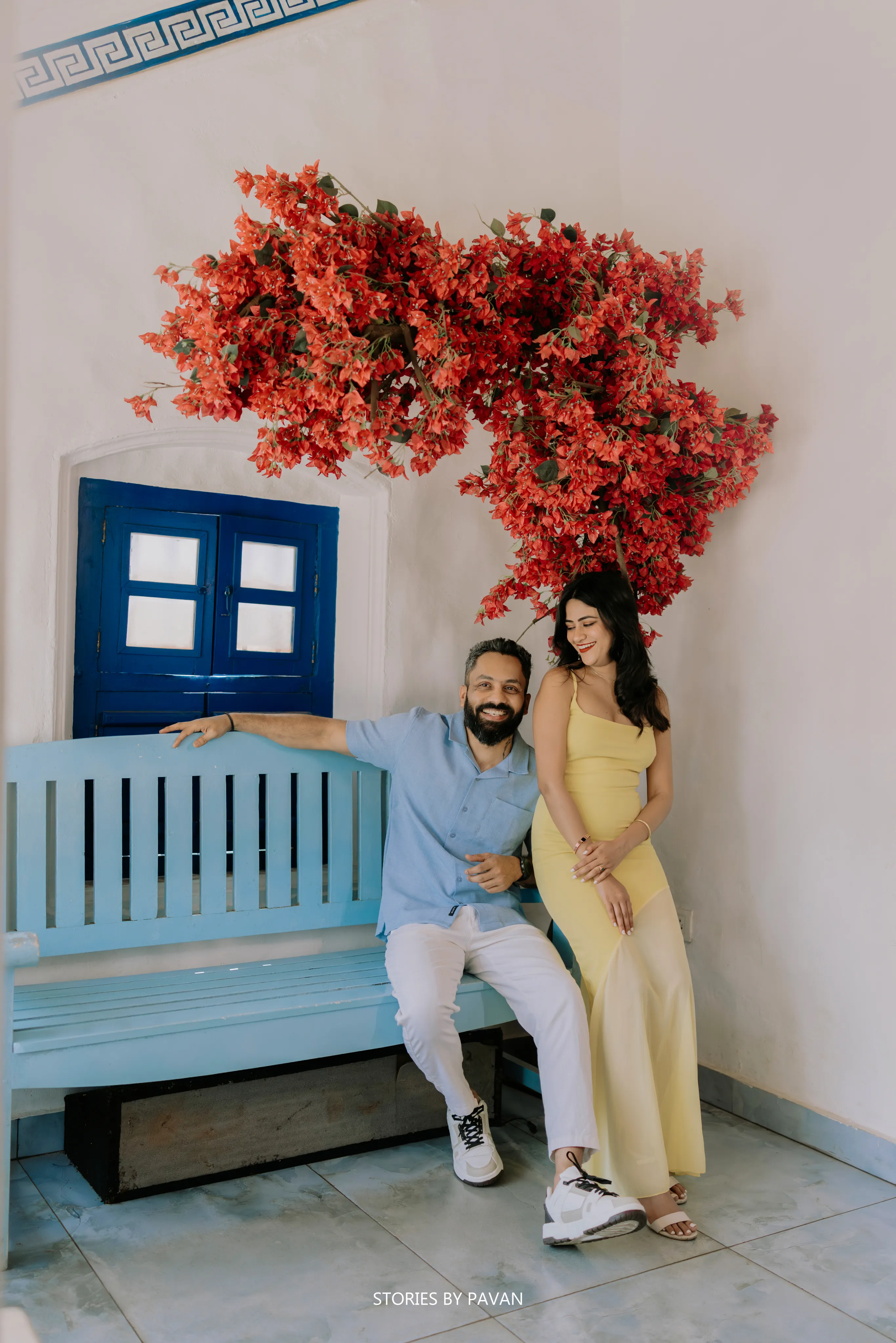 Couple sitting together on a blue bench beneath red bougainvillea flowers and a blue window